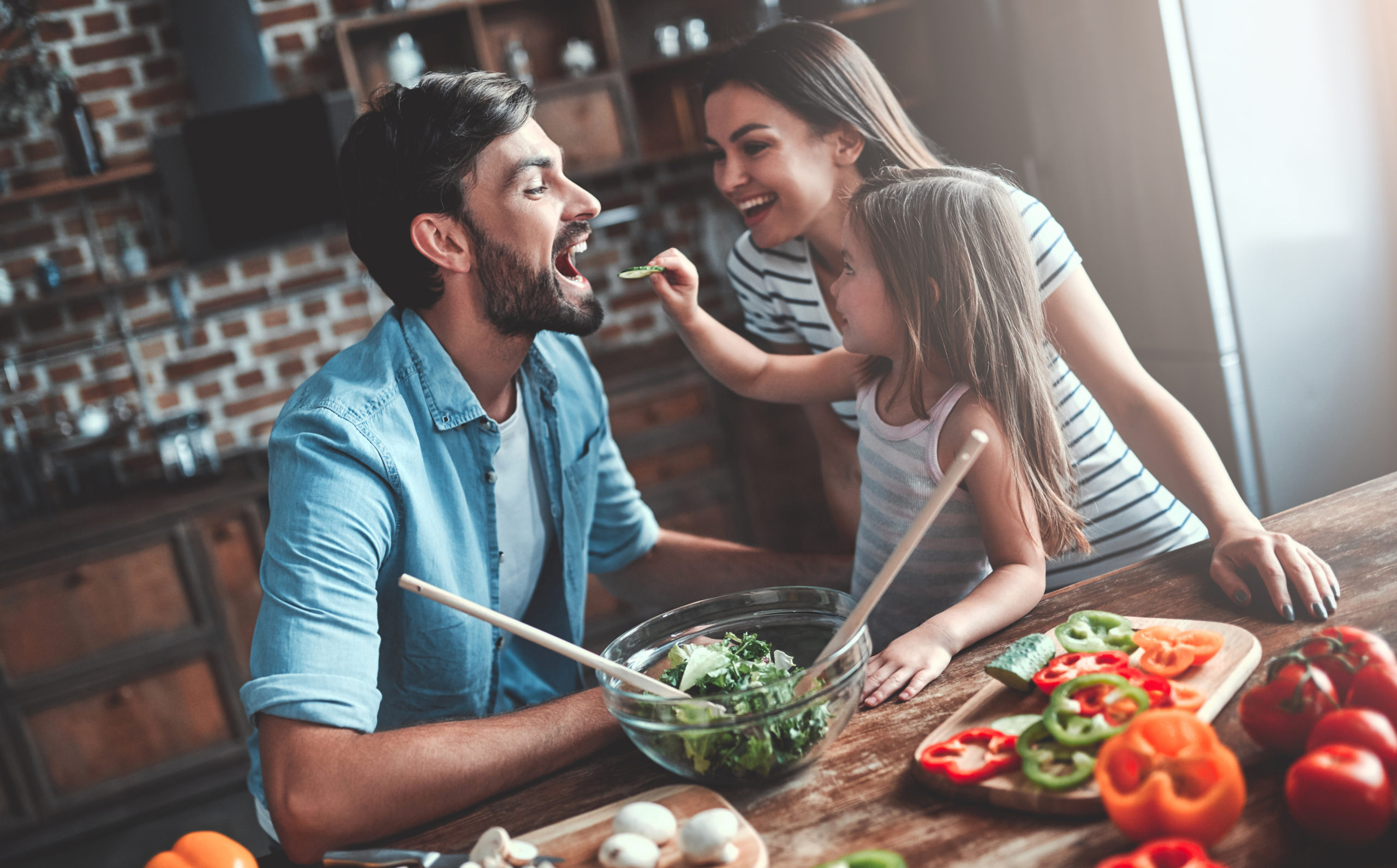 Mom, dad and daughter are cooking on kitchen. Happy family concept. Handsome man, attractive young woman and their cute little daughter are making salad together. Healthy lifestyle.