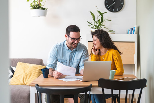 Happy husband and wife read good news online at laptop, smiling man holding documents receiving positive decision from bank