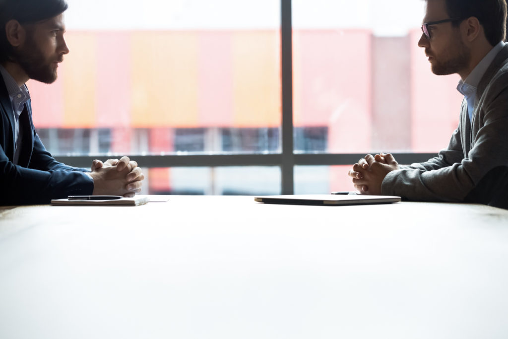 Side view two confident serious businessmen with tensed nerves sit at boardroom desk looking at each other, rivalry bad attitude, confrontation during difficult negotiation, leadership tussle concept