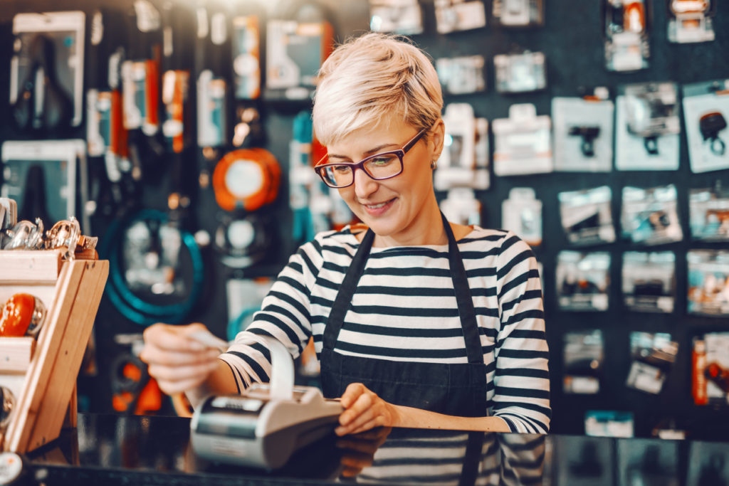 Smiling Caucasian female worker with short blonde hair and eyeglasses using cash register while standing in bicycle store.