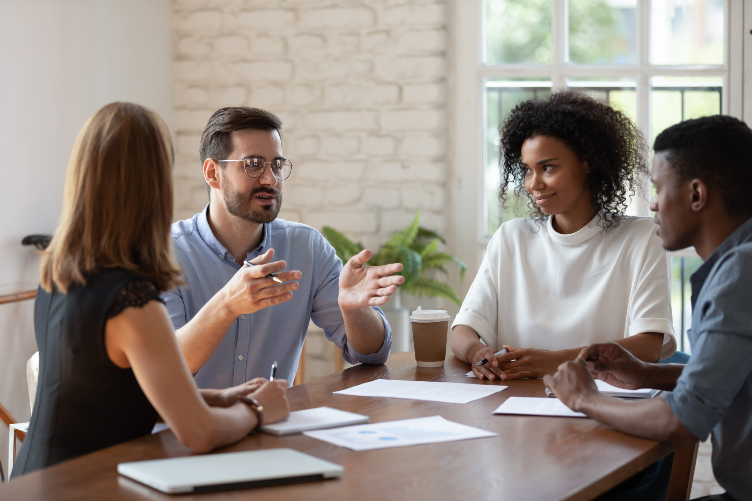 Diverse business partners brainstorming, discussing project at meeting, sitting at table in boardroom, colleagues listening to businessman sharing ideas, confident coach mentor training staff