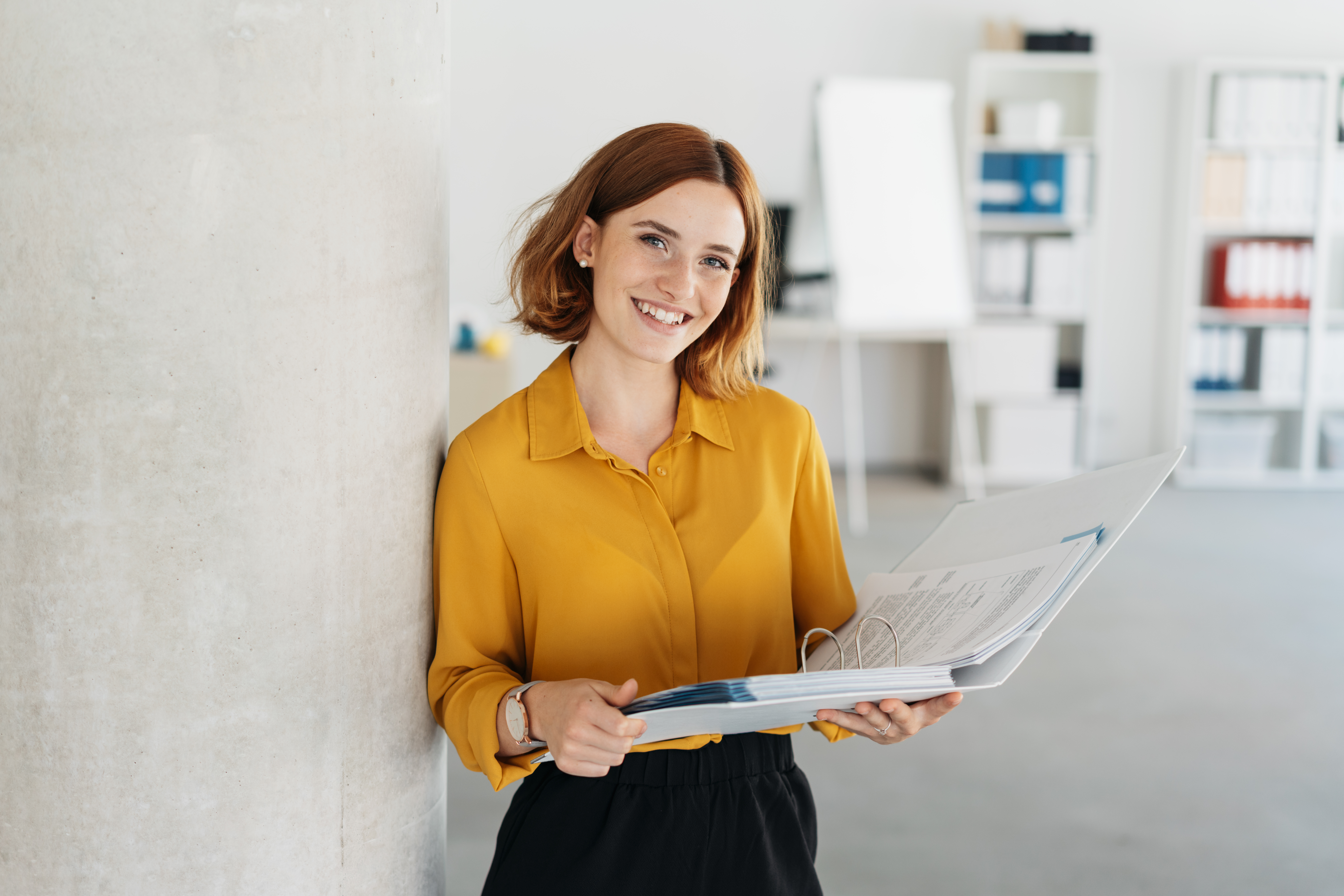 Attractive young office worker holding a large open binder as she looks at the camera with a sweet friendly smile