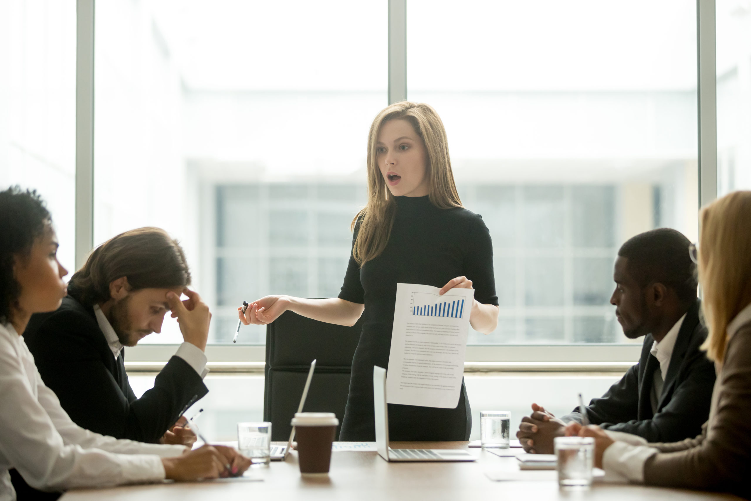 Dissatisfied angry female executive scolding african employee for bad work at diverse group meeting, white woman boss reprimanding black subordinate for poor financial result at office team briefing