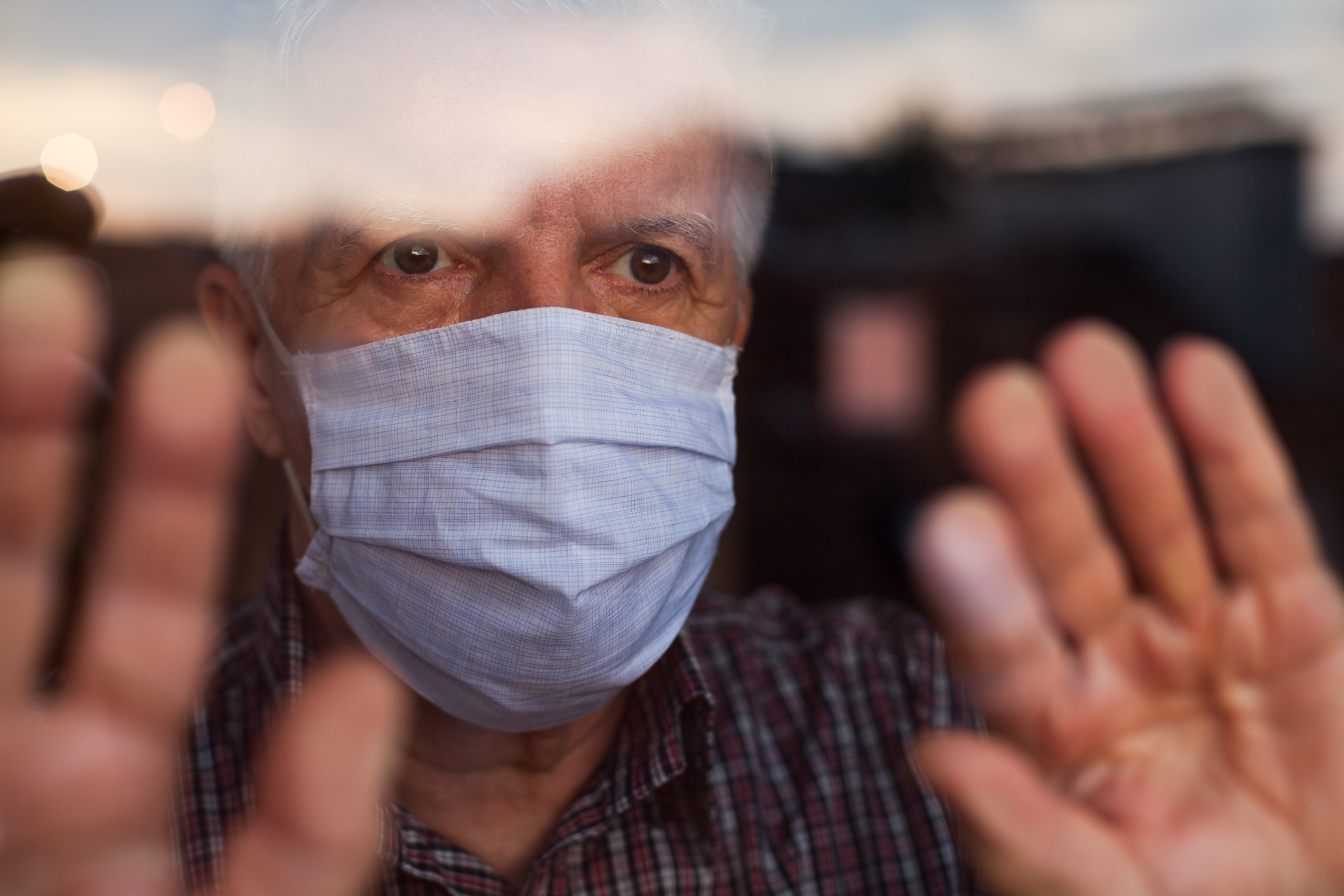 Elderly caucasian man wearing hand made protective face mask, in nursing care home, looking outside window, sadness, stress & hope in his eyes, self isolation due to global COVID-19 Coronavirus pandemic