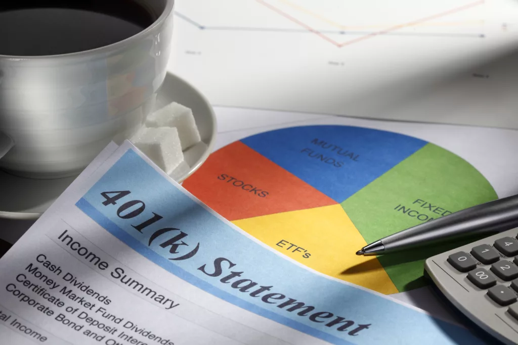 Close up of a 401(k) statement. The statement is surrounded by a cup of coffee, a pie chart of personal finances, a calculator and a pen with a soft focus on a graph in the background.  This image portrays the process of calculating personal finances and planning for retirement.