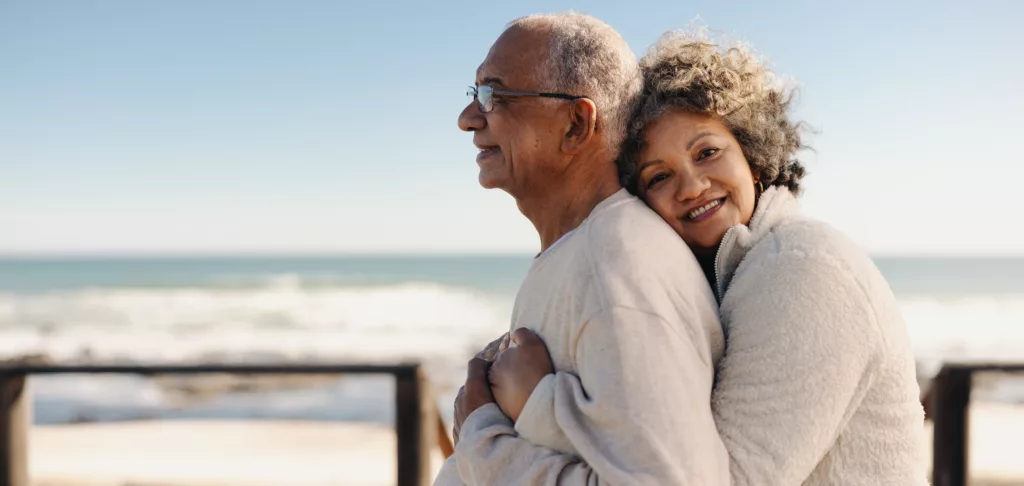 Romantic senior woman smiling at the camera while embracing her husband by the ocean. Affectionate elderly couple enjoying spending some quality time together after retirement.