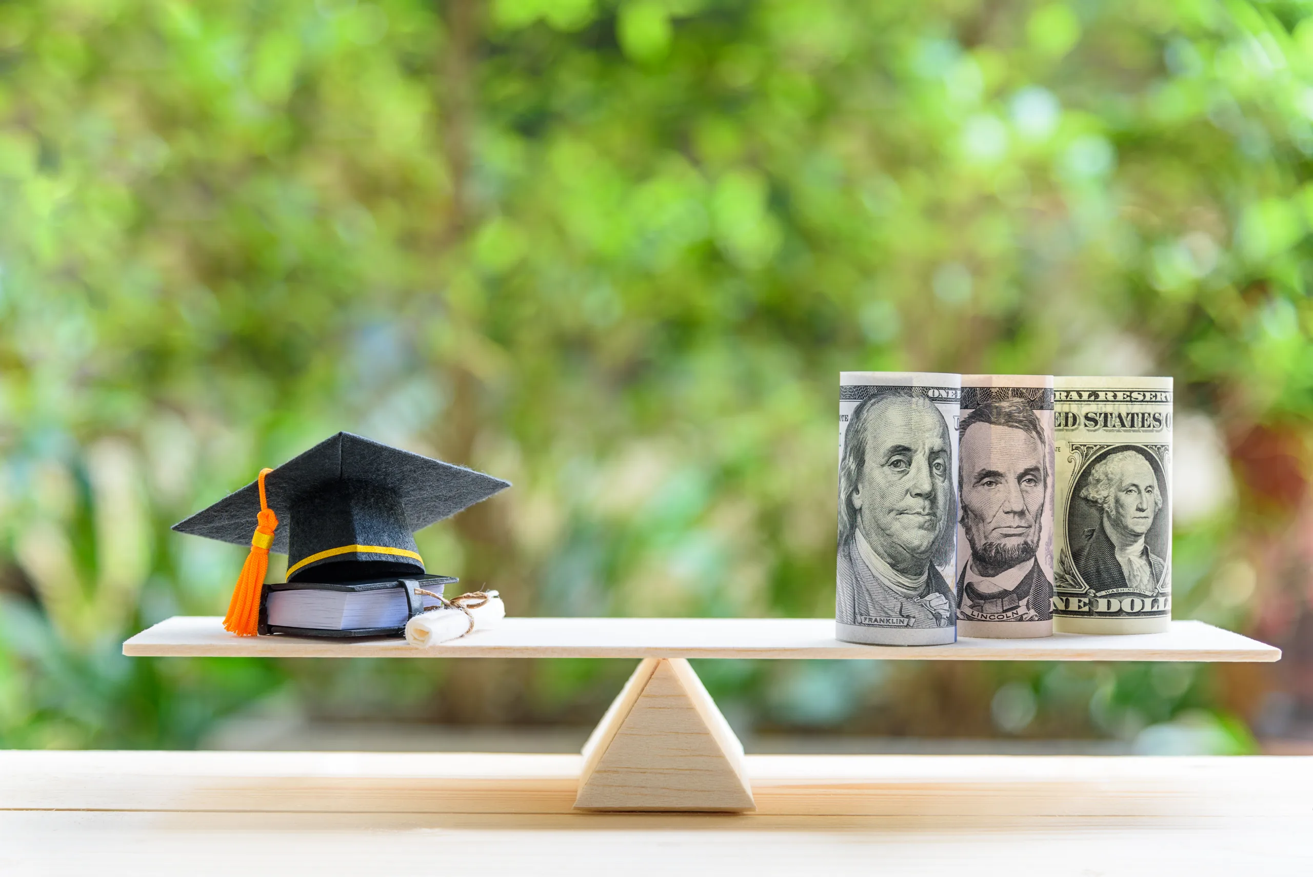 Money cost saving for goal and success in school, education concept : US USD dollar notes or cash, graduation cap, a text book, a certificate / diploma on basic wooden balance scale. Green background.
