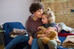 Young woman giving hug to her cute little son with brown soft teddybear while both sitting on sleeping place prepared for refugees