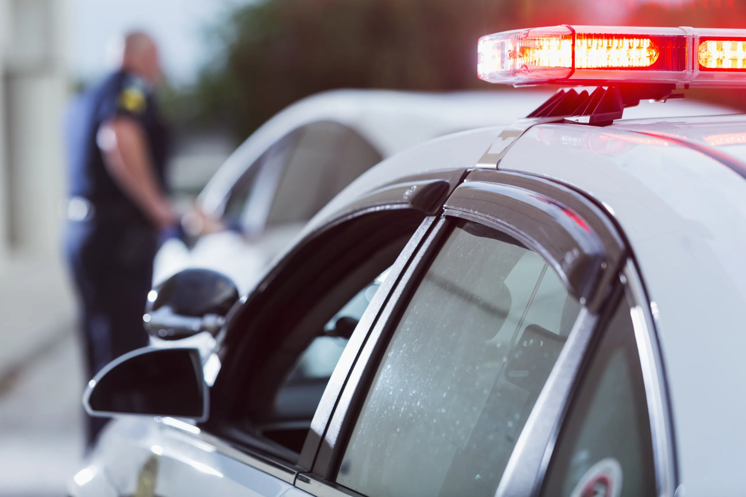 A police officer stands next to a car he pulled over for speeding,  reaching for the driver's ID.  The focus is on the police car in the foreground with lights on.  The policeman is out of focus in the background and unrecognizable.