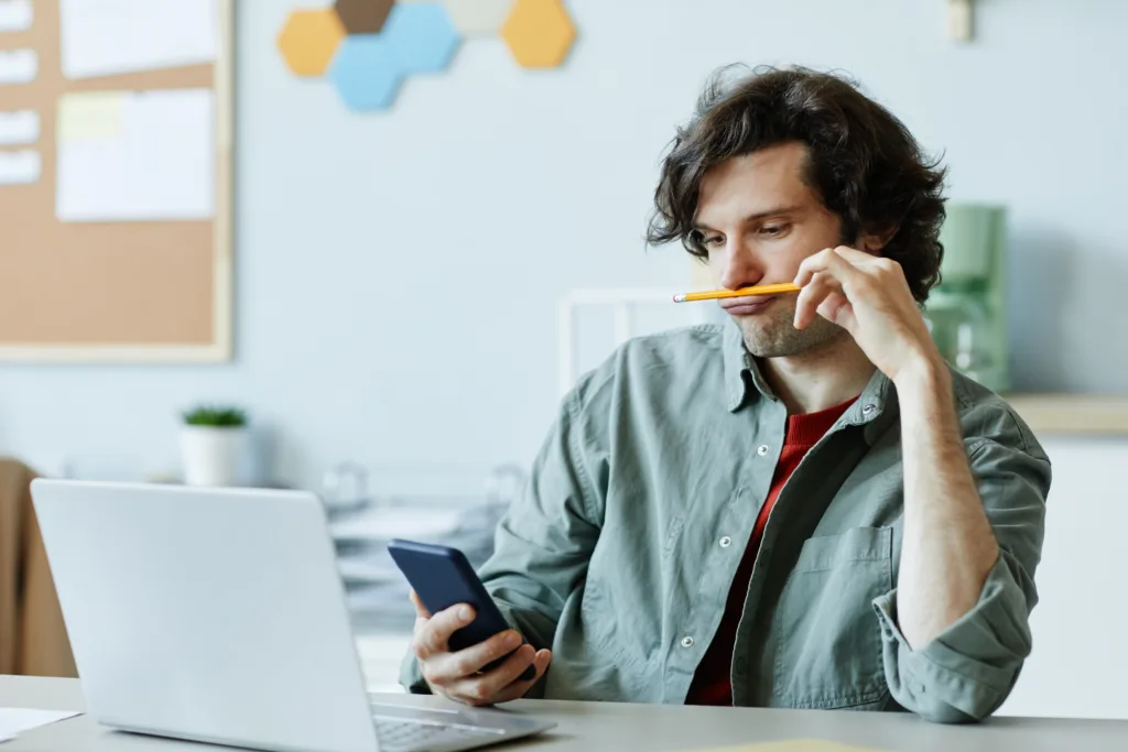 Portrait of Caucasian young man playing with pencil and using phone at workplace suffering from boredom and procrastination